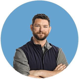 A headshot of Philip Martin, Coinbase CSO, smiling in a grey button-up and navy quarter-zip vest